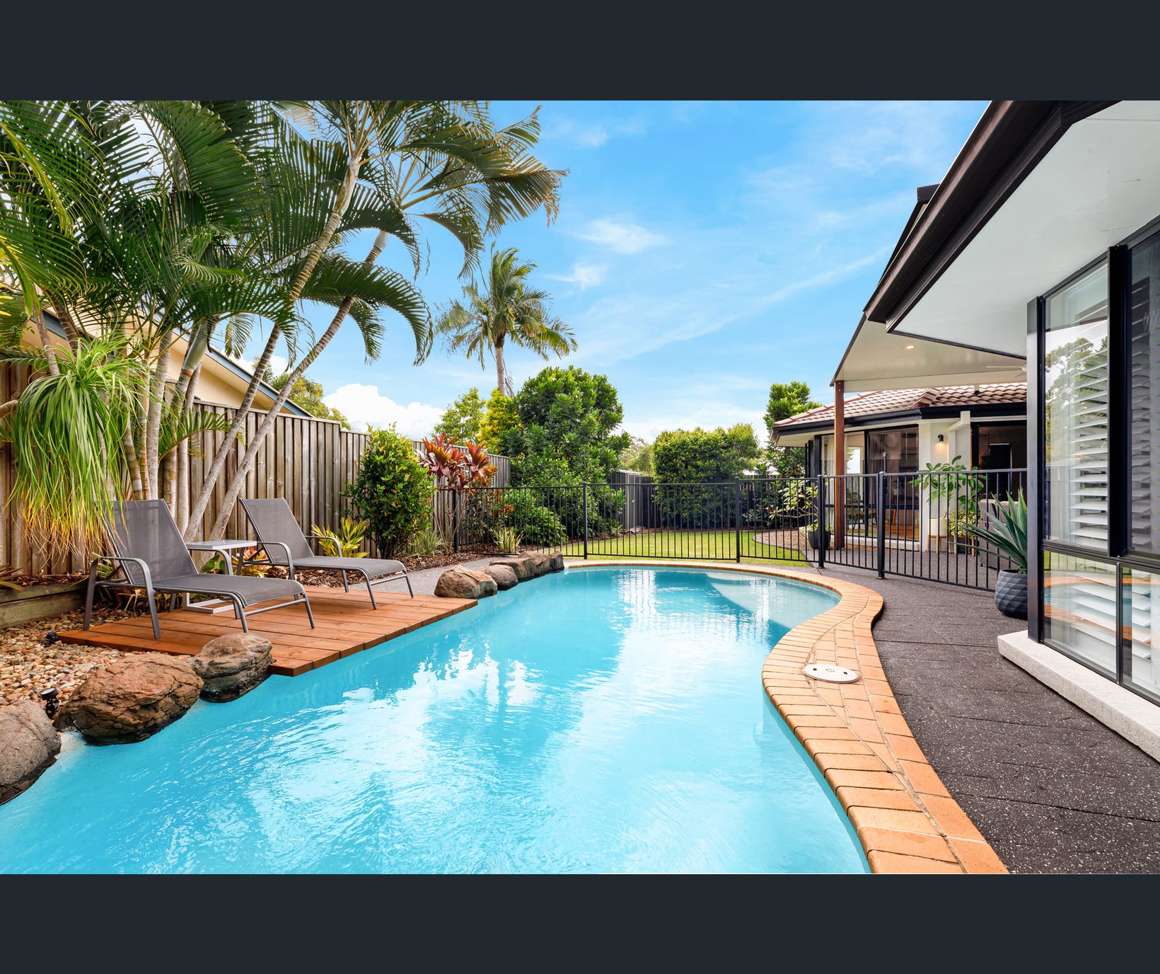 A backyard swimming pool with clear blue water, surrounded by tropical plants and palm trees. Two gray lounge chairs sit on a small wooden deck beside natural-looking stones. A black metal safety fence encloses part of the pool area near a modern house with large windows and a covered patio. The sky is bright and clear, creating a relaxing outdoor atmosphere.